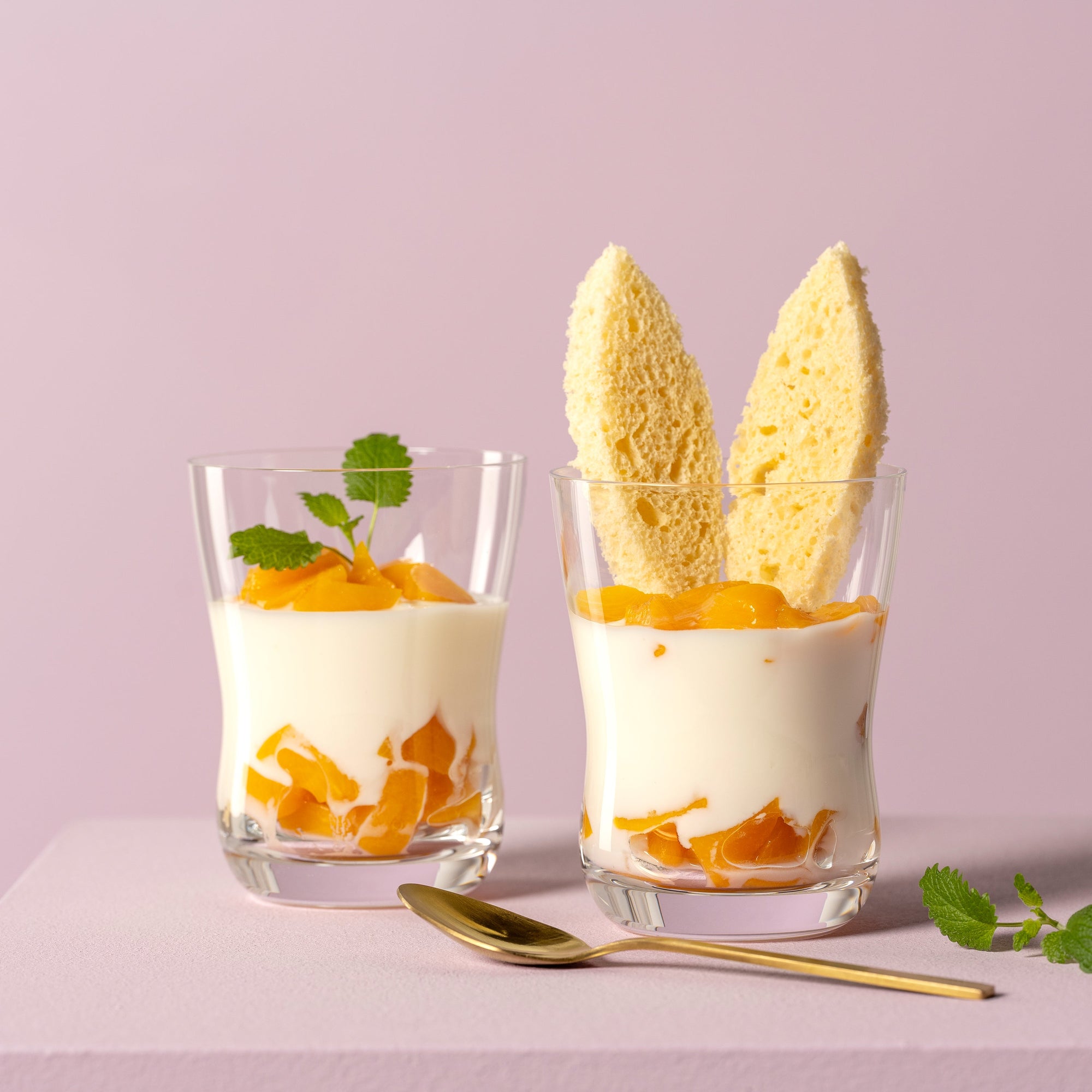 Zwei Dessertgläser mit Joghurt, Pfirsichstücken und Biskuit auf rosa Untergrund, daneben ein goldener Löffel.