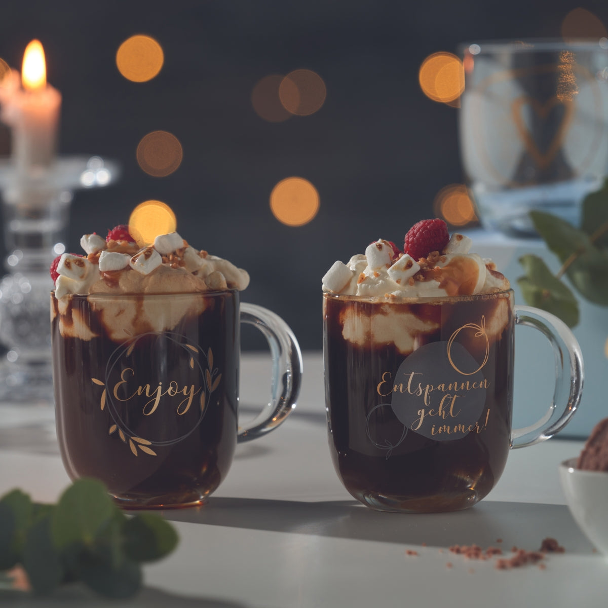 Zwei Tassen mit heißer Schokolade, Sahne, Marshmallows und goldenen Schriftzügen.