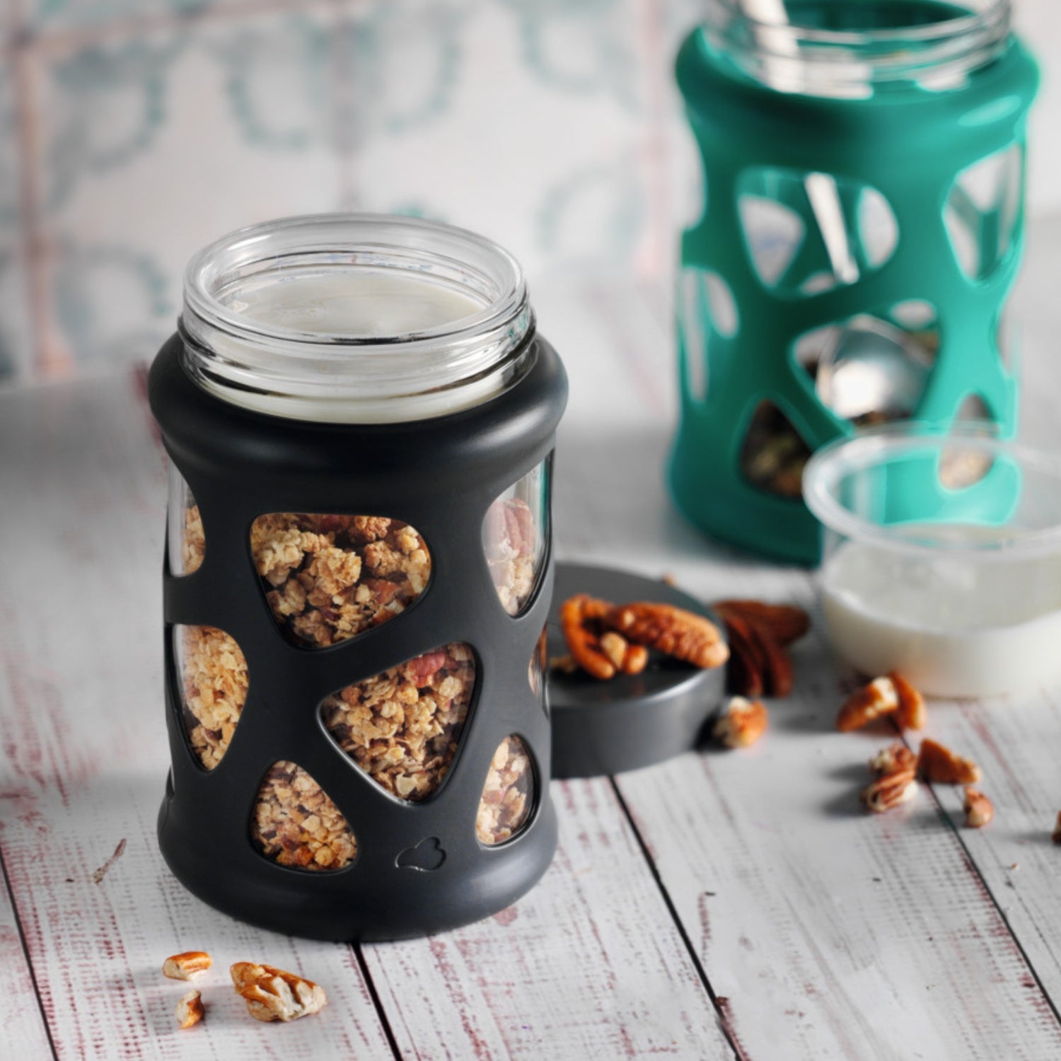 Knuspermüsli mit Joghurt im Glas mit schwarzer Silikonhülle – im Hintergrund ein weiteres Glas mit türkiser Hülle.