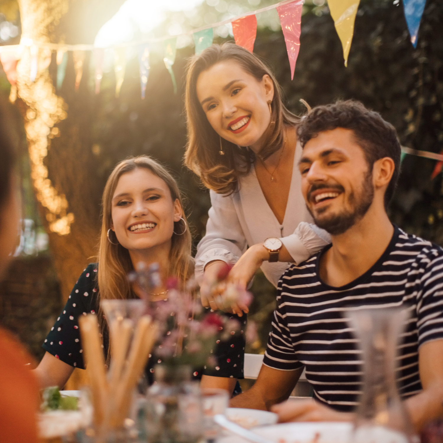 Drei junge Erwachsene lachen herzlich bei einem Sommerfest im Garten, im Hintergrund bunte Wimpel und warmes Sonnenlicht in gemütlicher Atmosphäre.