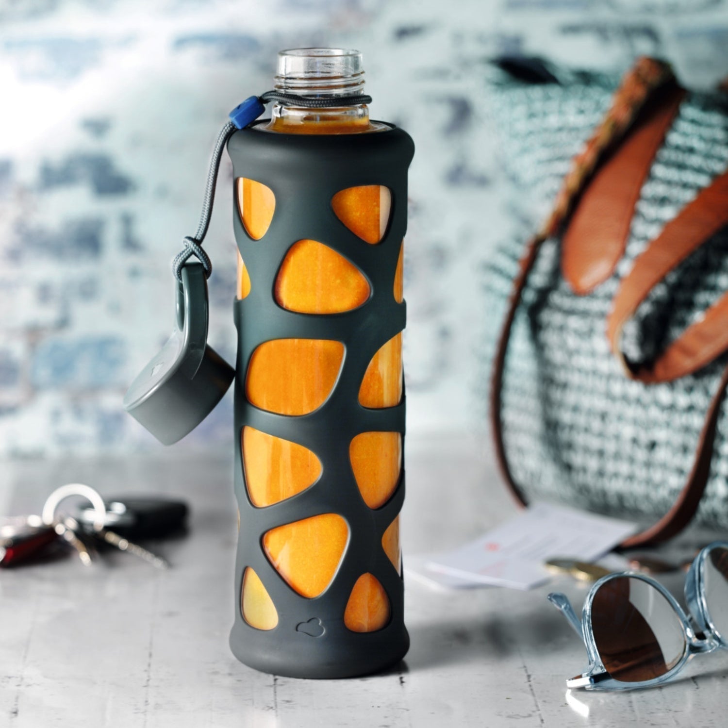 Glasflasche mit orangefarbenem Saft und schwarzer Hülle vor Tasche