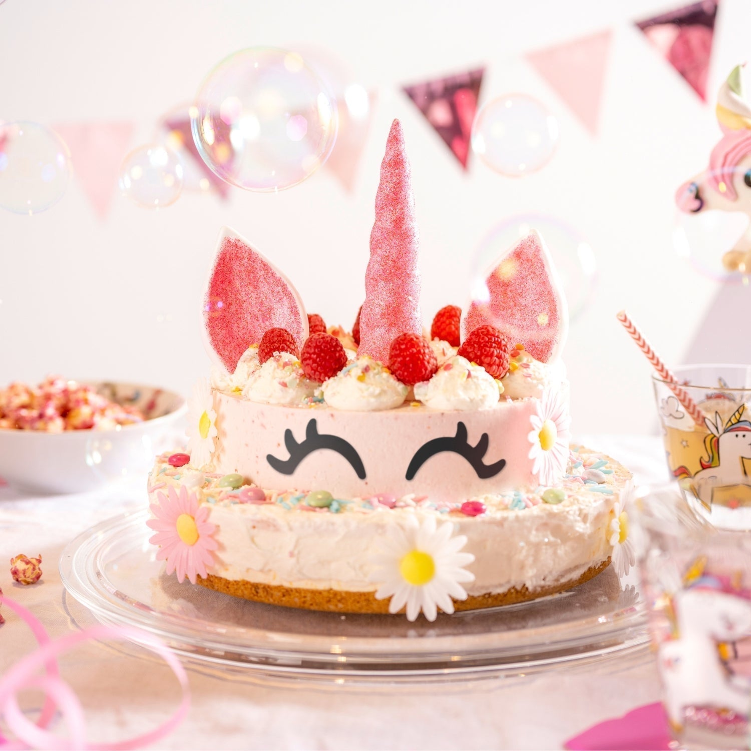 Einhorn-Torte mit pinkem Horn, rosa Ohren, Sahnetuffs, Himbeeren und bunten Streuseln auf einem Glasteller, dekoriert mit Blüten und Luftblasen im Hintergrund.