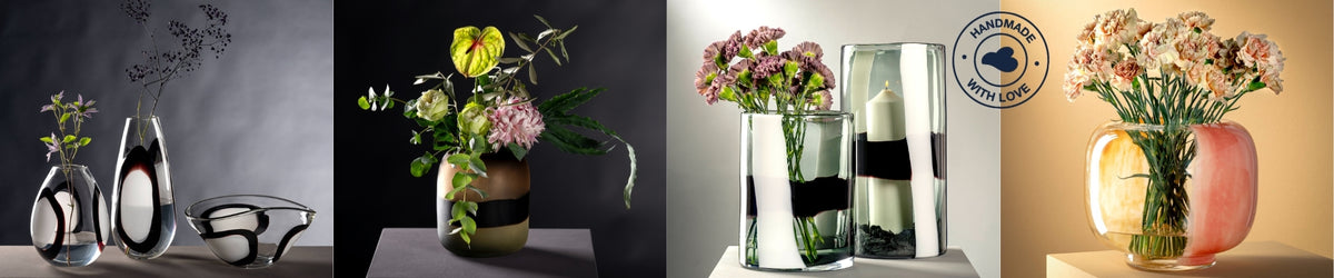 Eine stilvolle Komposition handgefertigter Glasobjekte mit unterschiedlichen Formen und Dekoren: Links organisch geformte Vasen und eine Schale mit schwarzer und weißer Musterung, in der Mitte eine goldfarbene Vase mit kunstvollem Blumenarrangement, daneben zwei zylindrische Vasen mit schwarzen und weißen Farbstreifen – eine davon mit violetten Nelken, die andere mit einer Kerze. Ganz rechts eine rundliche Vase mit orange-rosafarbenem Verlauf und pastelligen Nelken. Das Qualitätssiegel „Handmade with Love“
