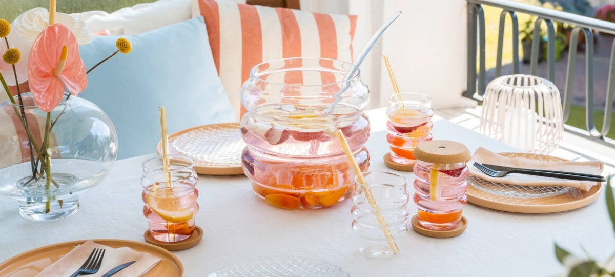 Sommerlich gedeckter Tisch auf dem Balkon mit der geschwungenen SOLE-Bowle aus Glas, passenden Trinkgläsern mit Obstbowle, Glastrinkhalmen und farblich abgestimmter Dekoration in Rosa, Apricot und Naturtönen.