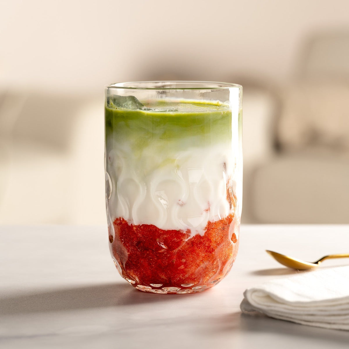 Farblich geschichteter Erdbeer-Matcha-Drink mit Milch in einem strukturierten Trinkglas von LEONARDO, auf einem hellen Tisch mit goldfarbenem Löffel und Stoffserviette.