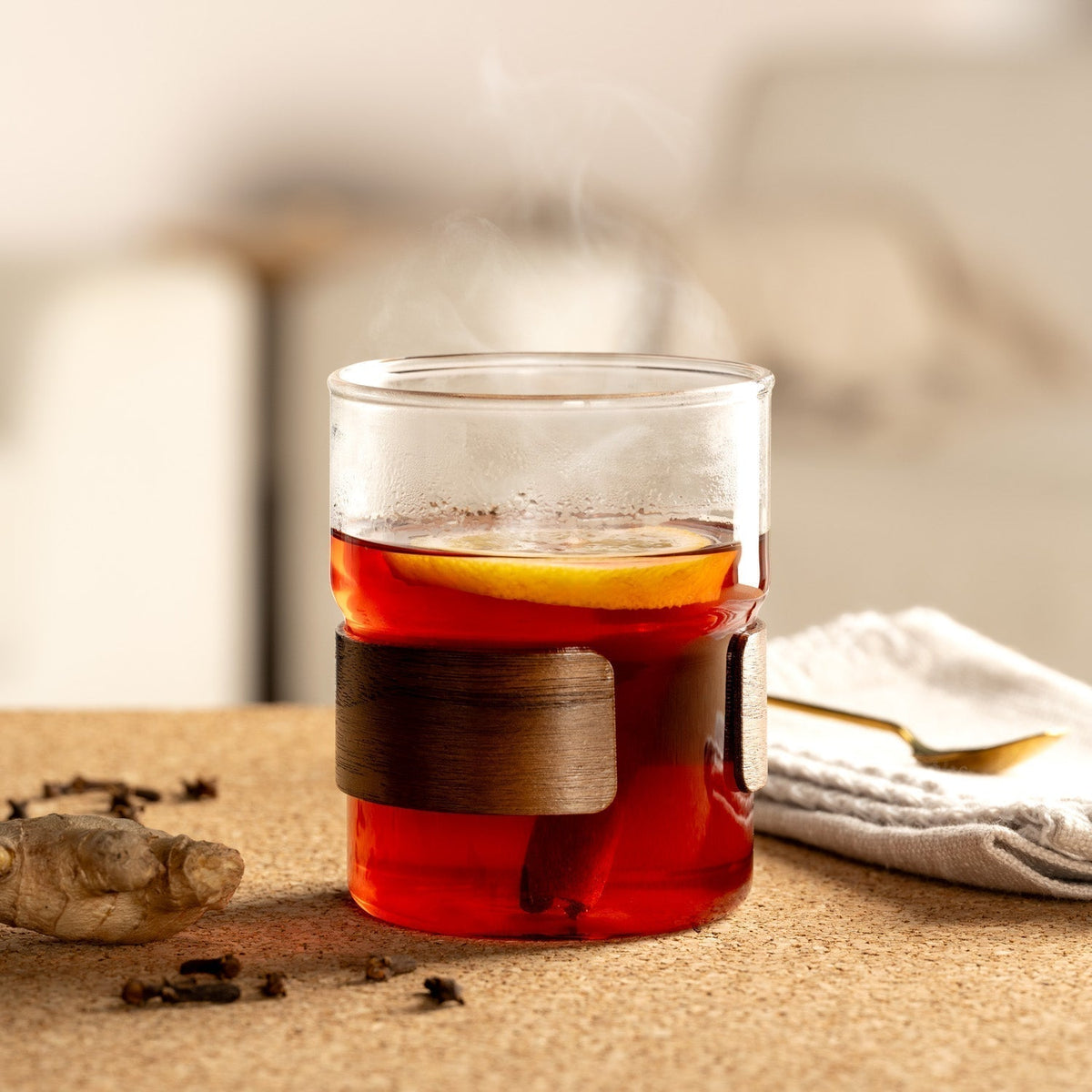 Ein dekoratives Teeglas mit schwarzem Tee, Zitronenscheibe und Zimtstange, serviert auf einem Korkuntergrund. Im Vordergrund liegen Gewürznelken und ein Stück frischer Ingwer.