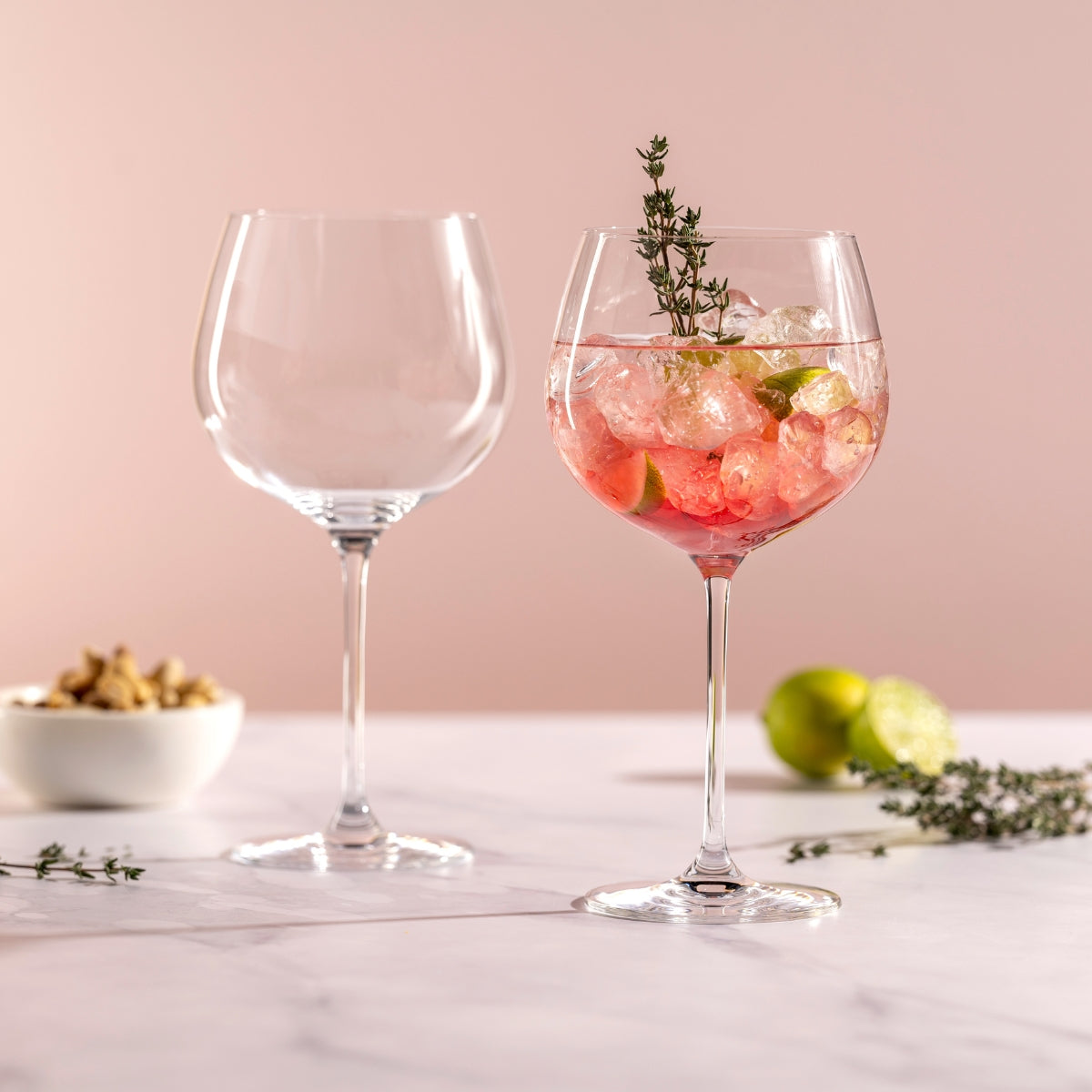 Bauchige Cocktailgläser mit Gin Tonic, Eis und Limette vor rosa Hintergrund