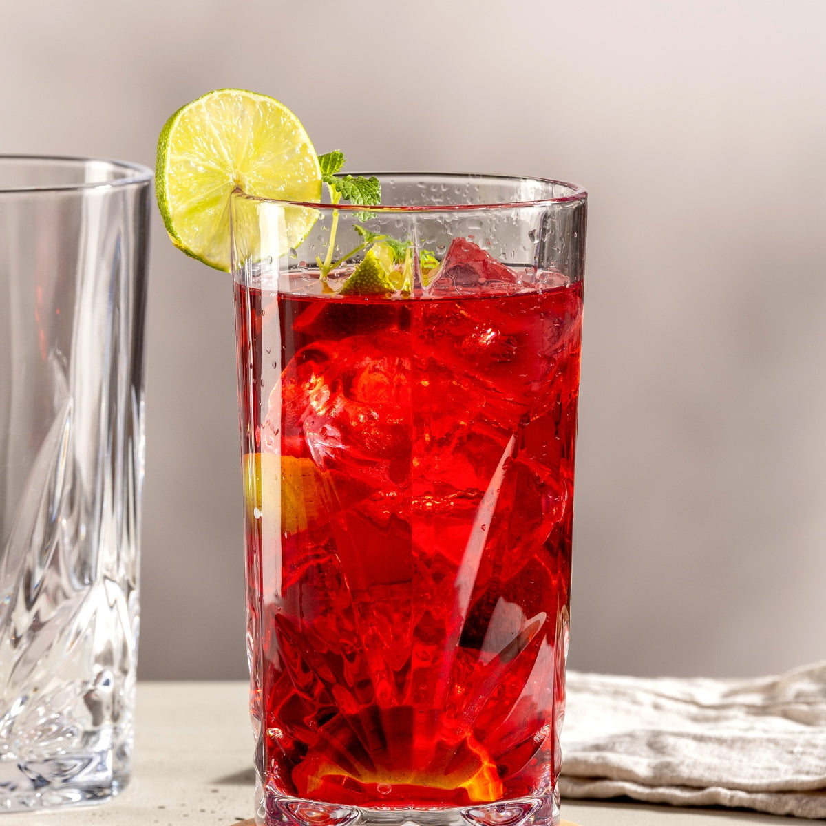 In einem eleganten Longdrinkglas mit Facettenschliff leuchtet ein roter Cocktail mit Eiswürfeln, frischer Limette und Minze