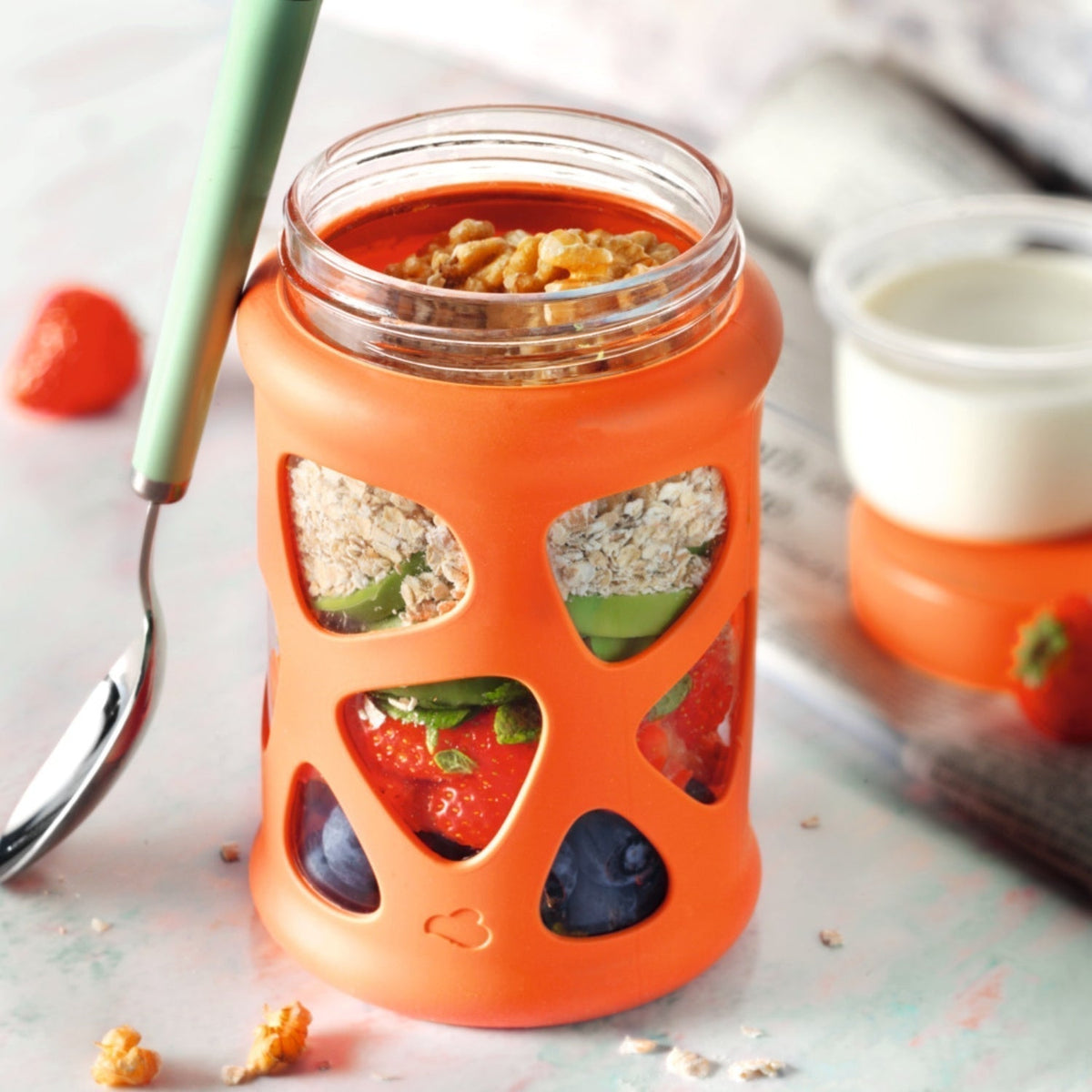 Schichtglas mit Haferflocken, Früchten und Nüssen in orangener Silikonhülle – daneben ein Becher Joghurt.