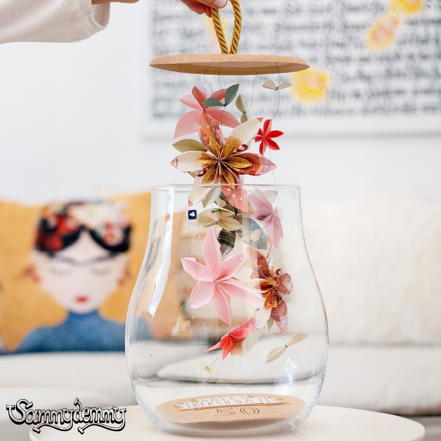 Eine handgefertigte Girlande aus Origami-Blumen und gefalteten Geldscheinen wird in eine bauchige Glasvase mit Holzdeckel gehängt