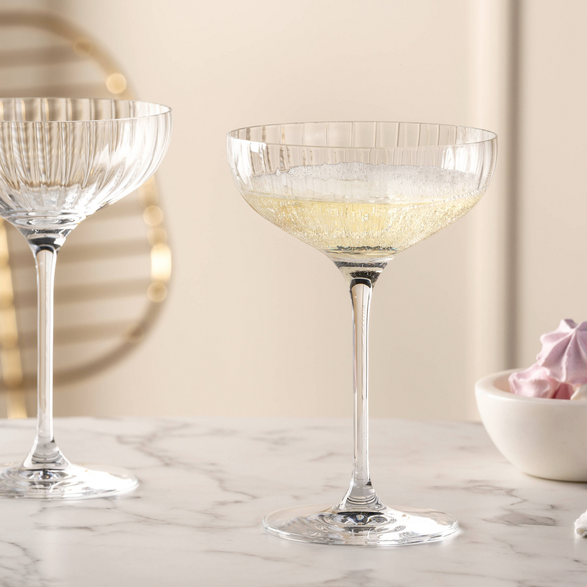 Ein mit Sekt gefülltes Champagnerschalen-Glas mit dezentem Rillendekor steht elegant auf einer Marmorplatte, im Hintergrund ist ein weiteres Glas leicht angeschnitten zu sehen.
