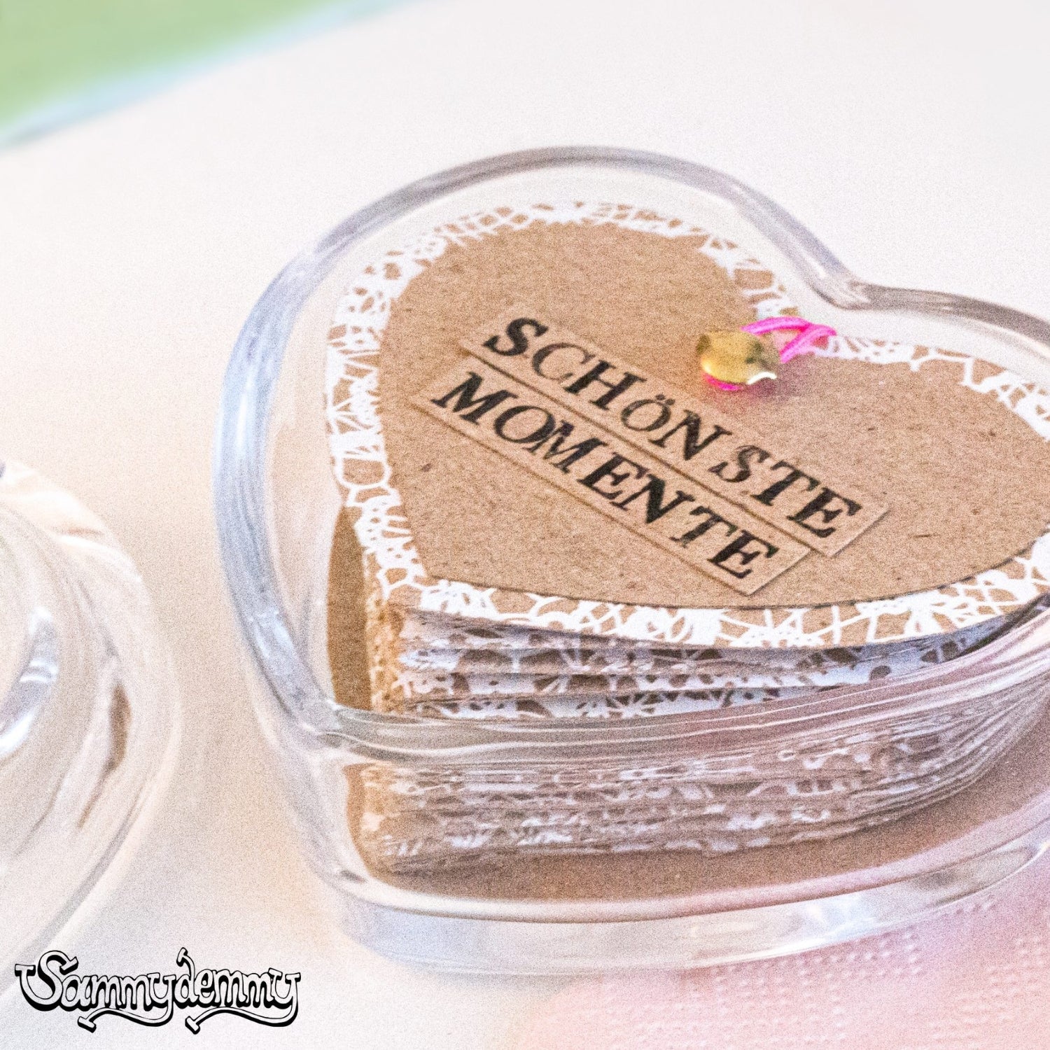Ein handgemachtes Erinnerungsalbum in Herzform mit der Aufschrift „Schönste Momente“ liegt in einer transparenten Herzdose. Die Herzseiten bestehen aus braunem Karton mit weißem Spitzenmuster und sind mit einem pinken Band zusammengeheftet.