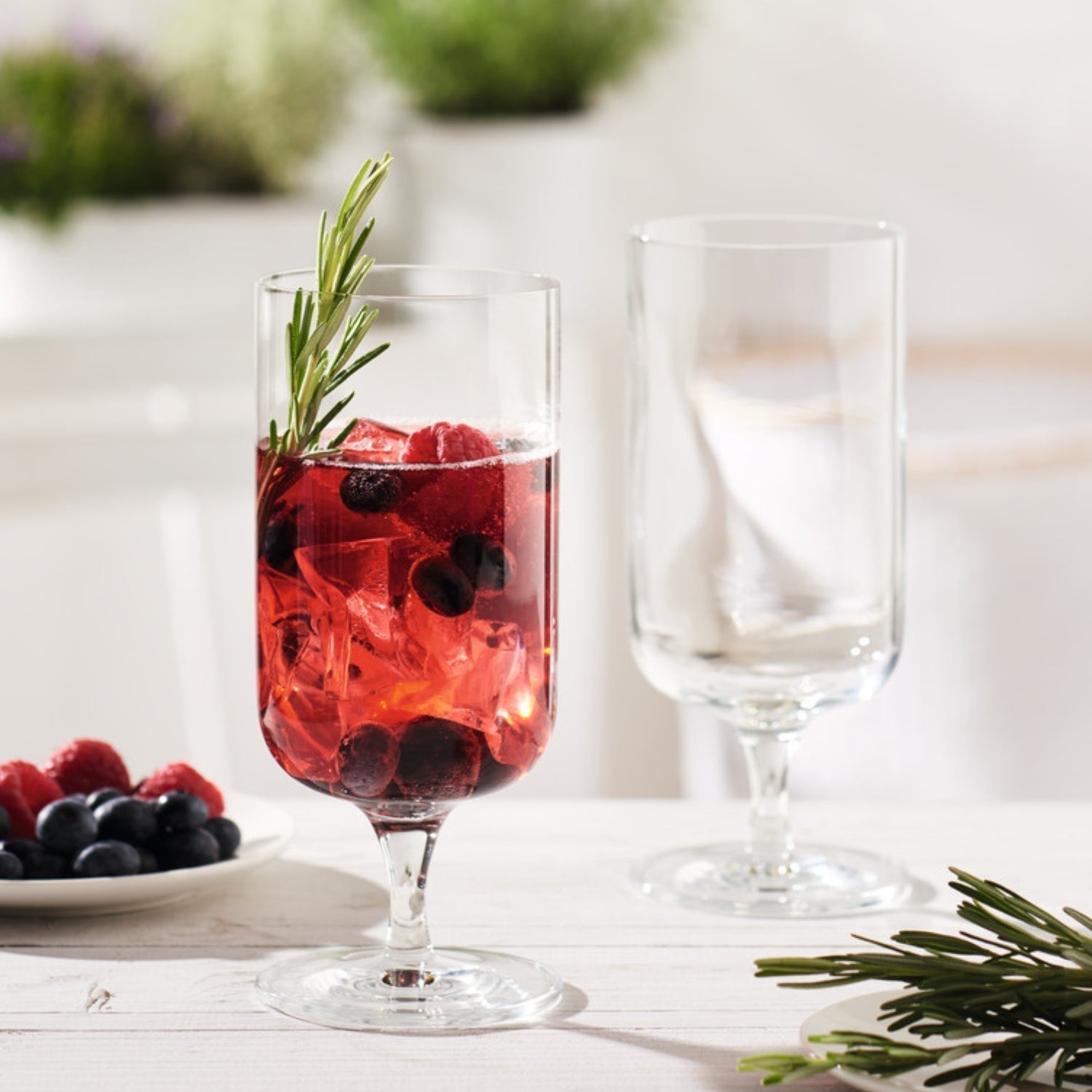Ein mit Beeren, Eiswürfeln und Rosmarin dekoriertes Getränk im eleganten Glas, daneben ein leeres Glas und frische Beeren.