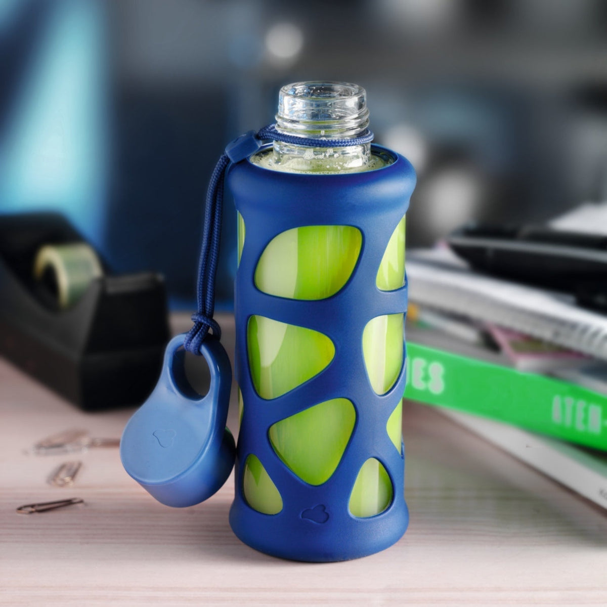 Kleine Glasflasche mit grünem Smoothie und blauer Hülle auf Schreibtisch