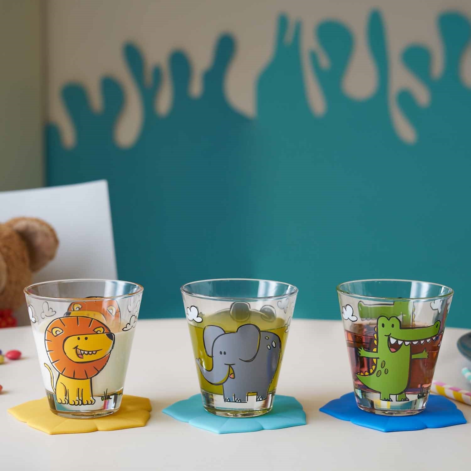 Drei Trinkgläser BAMBINI mit Tiermotiven auf farbigen Untersetzen, Tisch mit Deko im Hintergrund