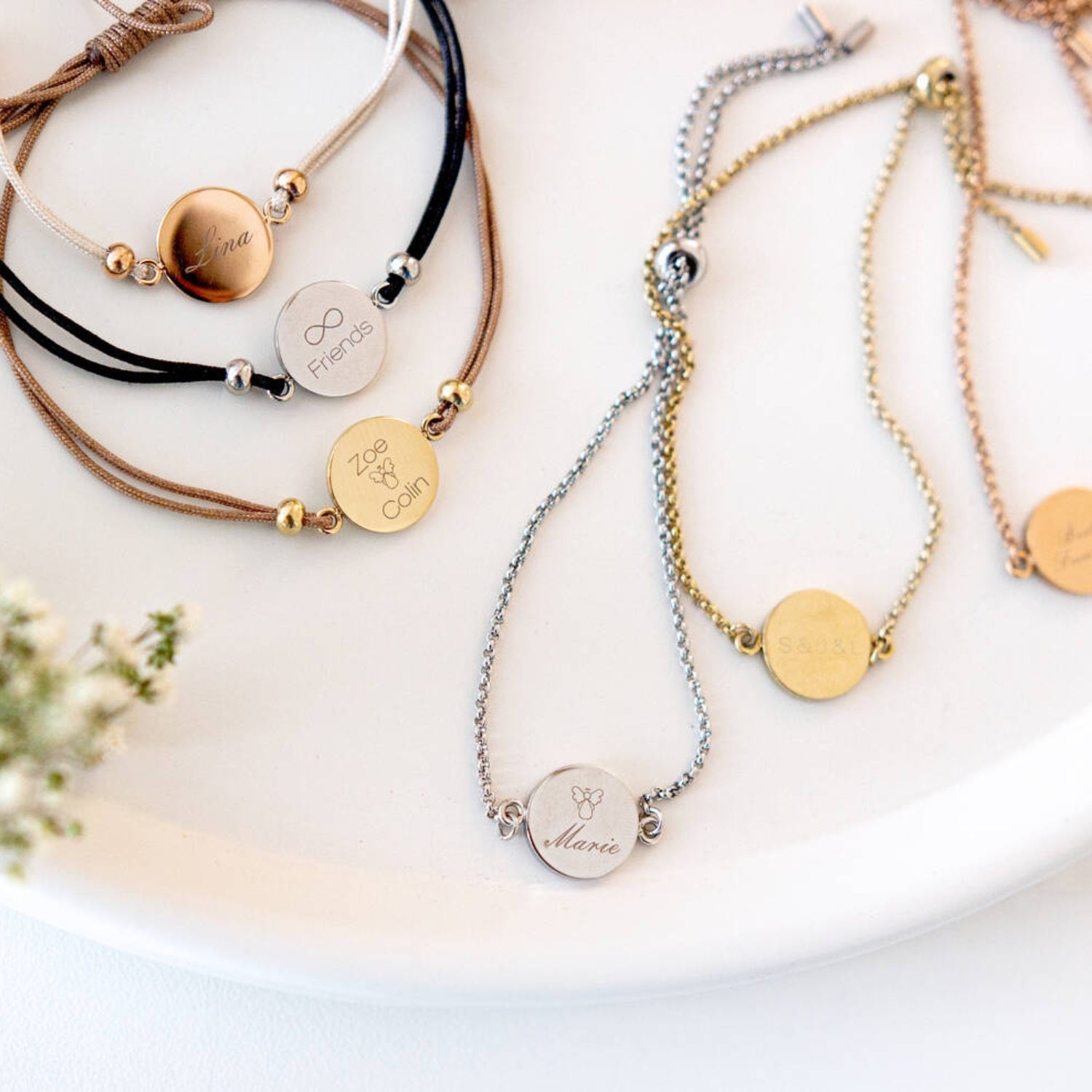 Mehrere Armbänder der Serie MARLA in Silber und Gold mit gravierten Plaketten auf weißer Oberfläche