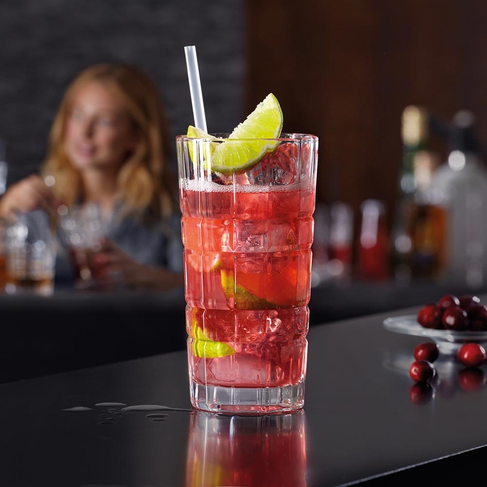 Longdrinkglas SPIRITII mit rotem Cocktail, Limettenscheibe und Strohhalm, Person im Hintergrund an der Bar