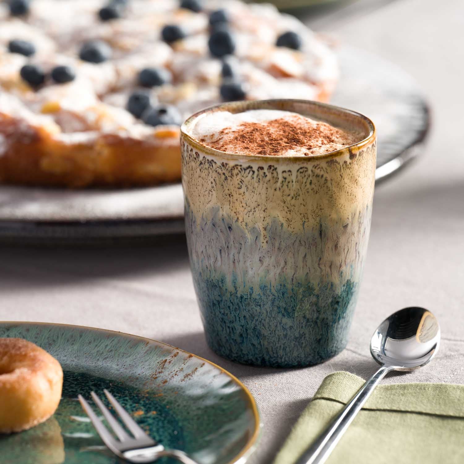 Keramikbecher MATERA mit Kaffee auf gedecktem Tisch, Gebäck im Hintergrund