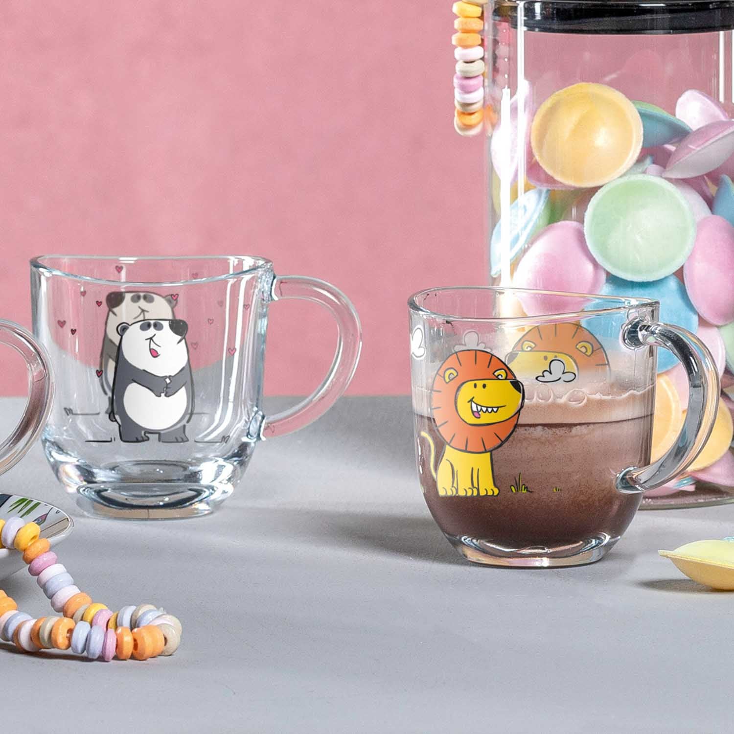 Tasse BAMBINI mit Löwen-Motiv neben Pandatasse auf grauem Tisch, rosa Hintergrund