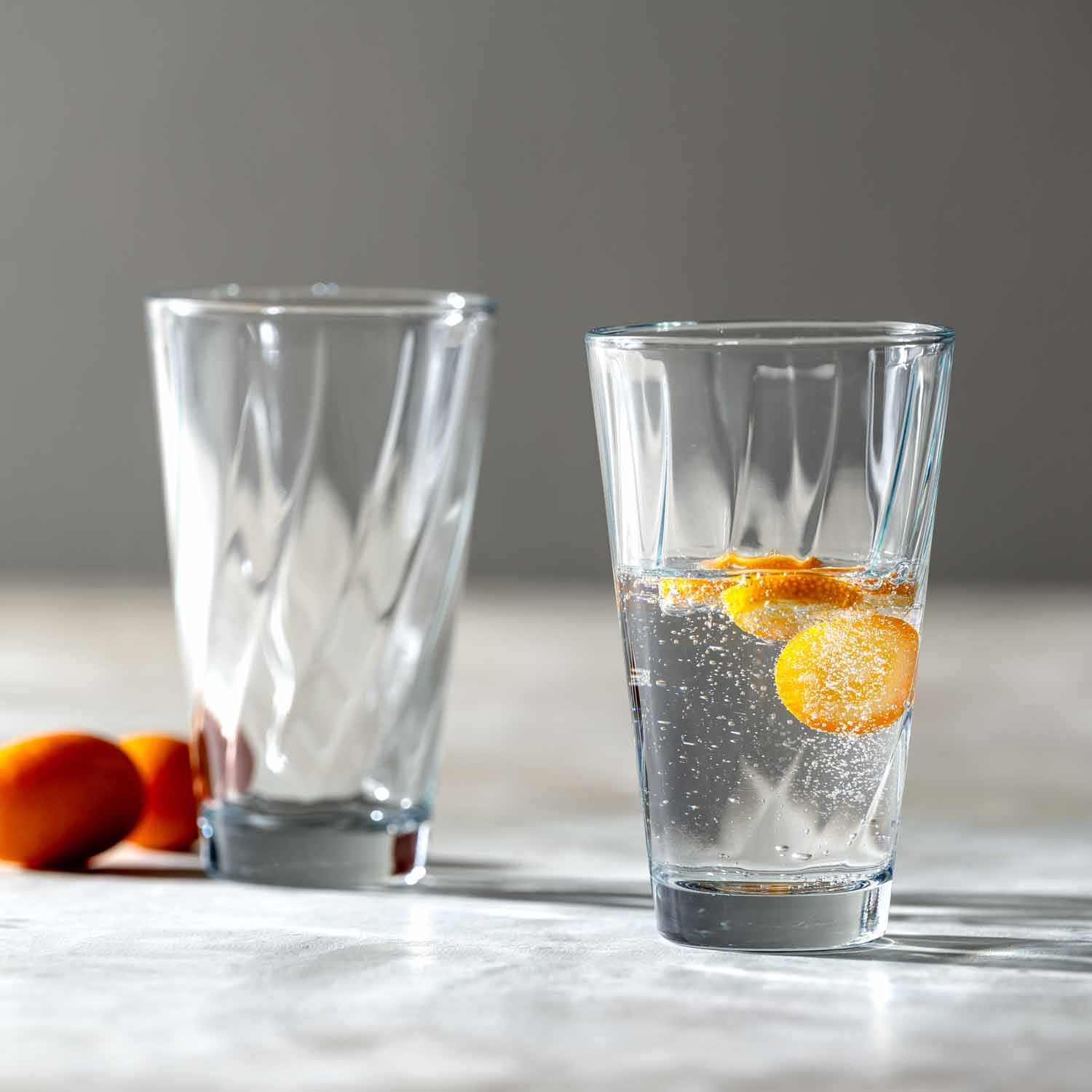 Zwei Gläser der Serie TWIST, eines leer, eines mit Wasser und Zitronenscheibe, auf heller Oberfläche