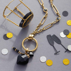 Halskette DELICA mit goldener Kette, schwarzem Herzanhänger und farbigen Glasperlen auf grauem Untergrund