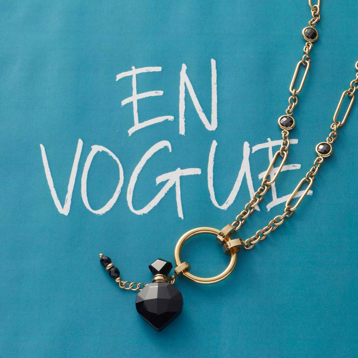 Halskette DELICA mit goldenem Kettenglied und schwarzem Herzanhänger auf türkisblauem Hintergrund mit "EN VOGUE" Schriftzug