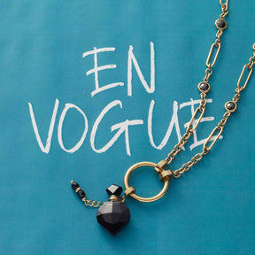 Halskette DELICA mit goldenem Kettenglied und schwarzem Herzanhänger auf türkisblauem Hintergrund mit "EN VOGUE" Schriftzug