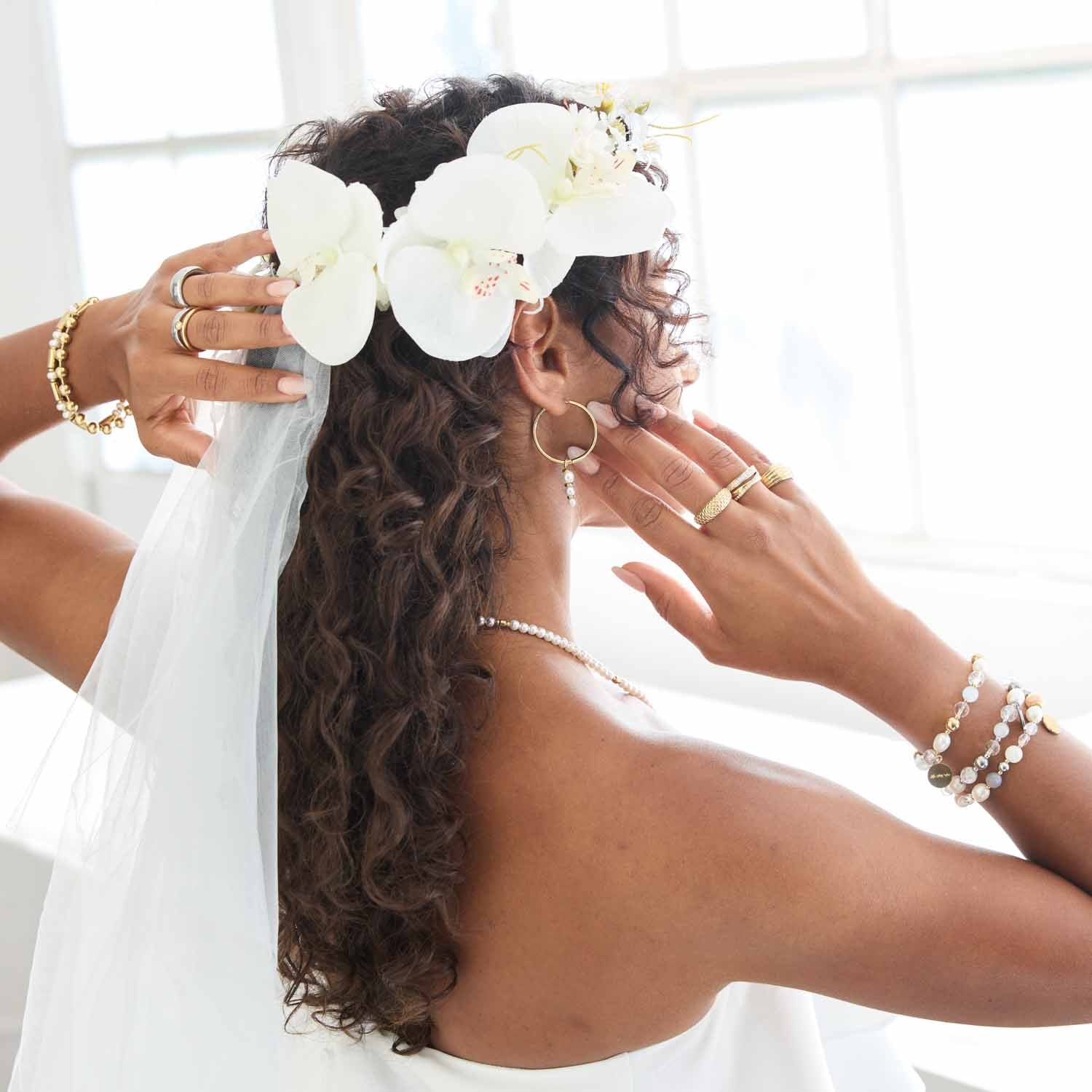 Frau von hinten mit weißen Orchideen im Haar, trägt goldene Armbänder und Ringe