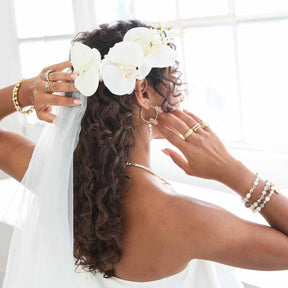 Frau von hinten mit weißen Orchideen im Haar, trägt goldene Armbänder und Ringe