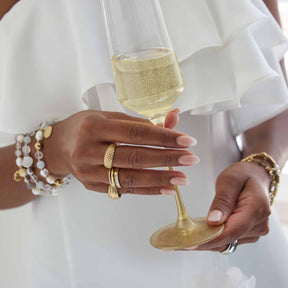 Frau in weißem Kleid mit Schmuck hält Weinglas, trägt Ring SARAH 19 gold