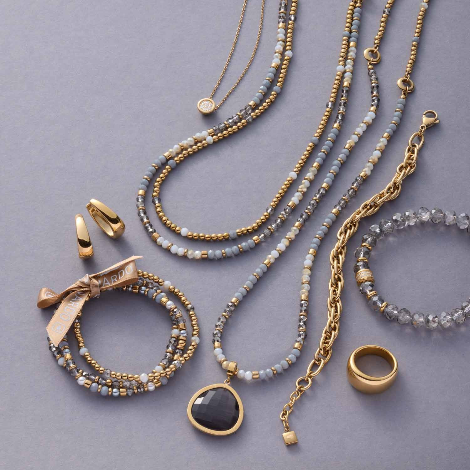 Creole ELODIE gold mit Perlenketten, Armbändern und Ringen auf grauem Untergrund