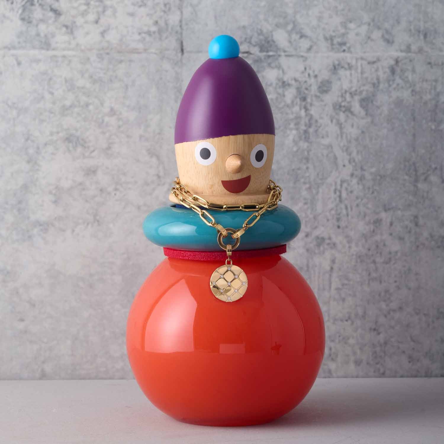 Bunte Holzfigur mit purpurner Kappe, türkisem Kragen und rotem Körper, trägt goldene Kette mit Anhänger