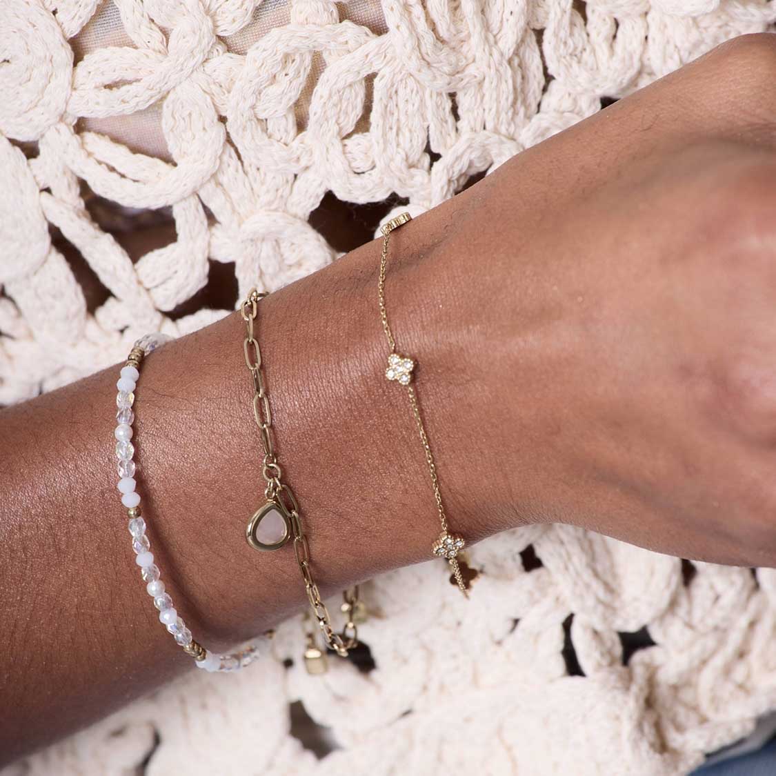 Hand mit mehreren goldenen Armbändern, weißem Perlenarmband und Spitzendetail im Hintergrund