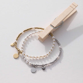 Drei Perlenbänder in Gold, Weiß und Silber mit Charms, neben Holzklammer