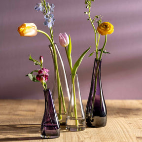 Drei violette Glasvasen mit Blumen auf Holztisch vor rosa Hintergrund