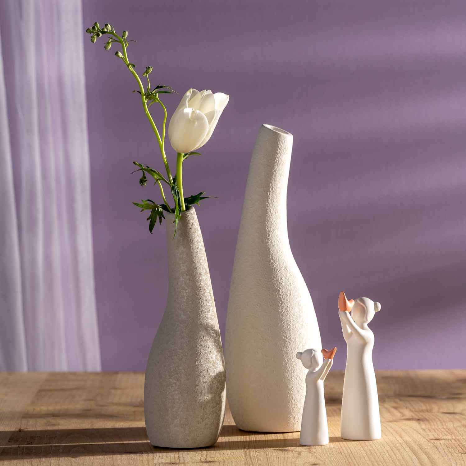 Weiße Keramikfiguren der Serie LUMINOSA auf Holzoberfläche vor violettem Hintergrund, daneben Vasen mit weißer Tulpe
