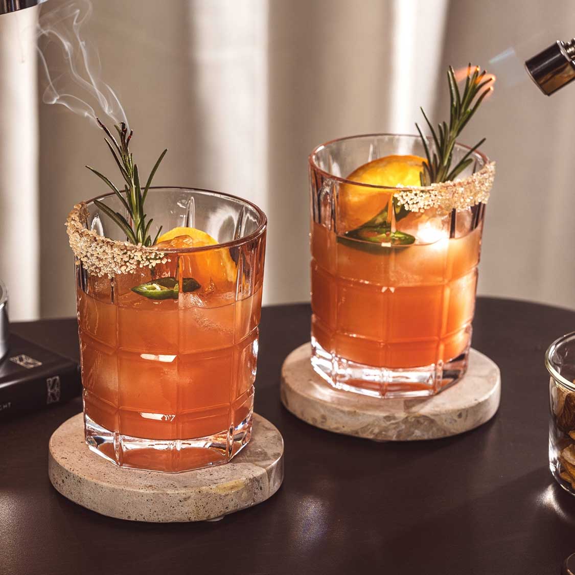 Zwei gefüllte Trinkgläser SPIRITII NOVA mit orangefarbenem Cocktail, Eiswürfeln, Rosmarin und Zitronenscheibe auf Untersetzer