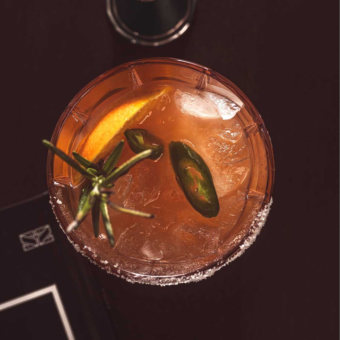Trinkglas SPIRITII NOVA mit orangebraunem Cocktail, Eiswürfeln und Jalapeño-Scheiben als Garnitur