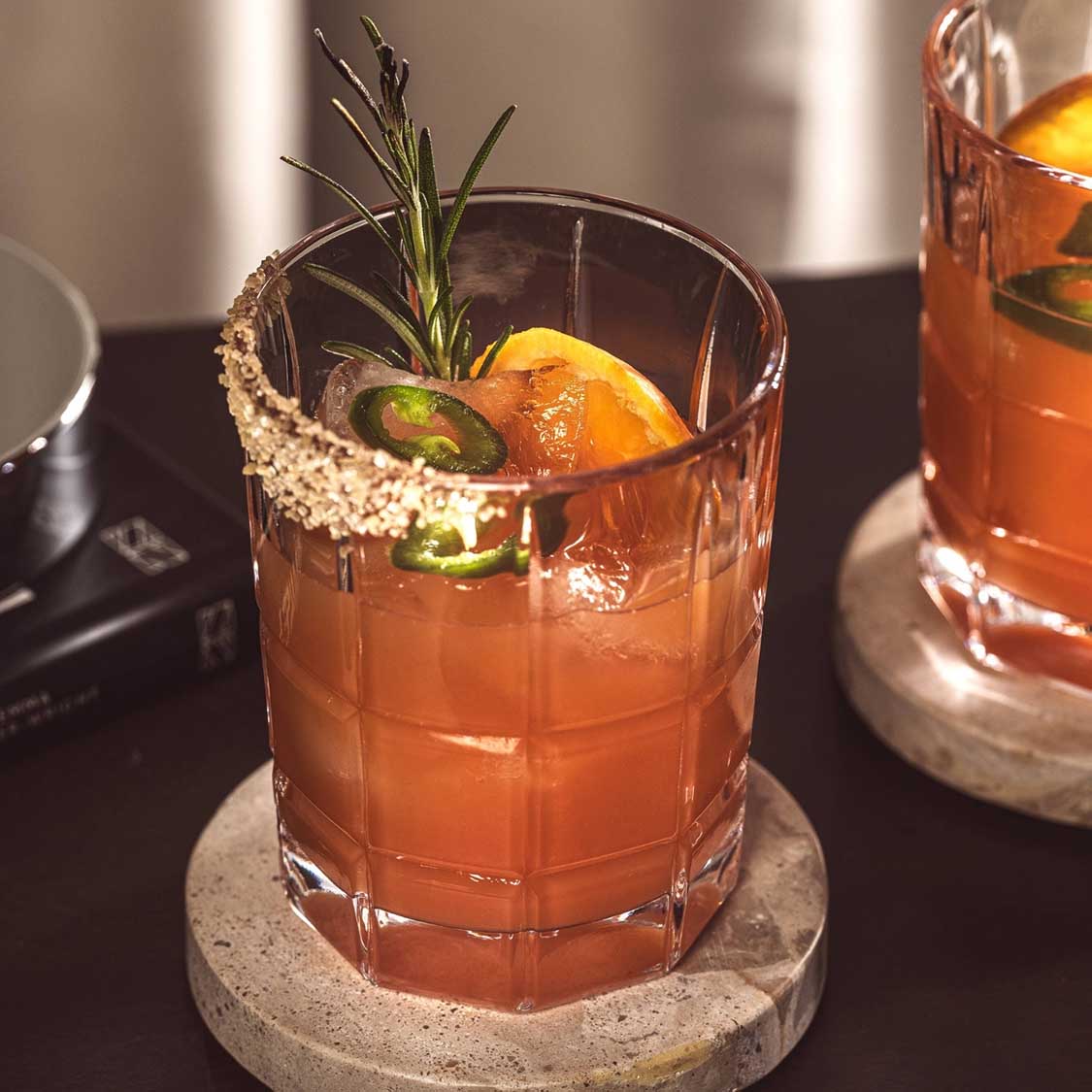 Trinkglas SPIRITII NOVA mit orangenem Cocktail, Rosmarin, Orangenscheibe und Jalapeño-Scheibe, auf Betonuntersetzer