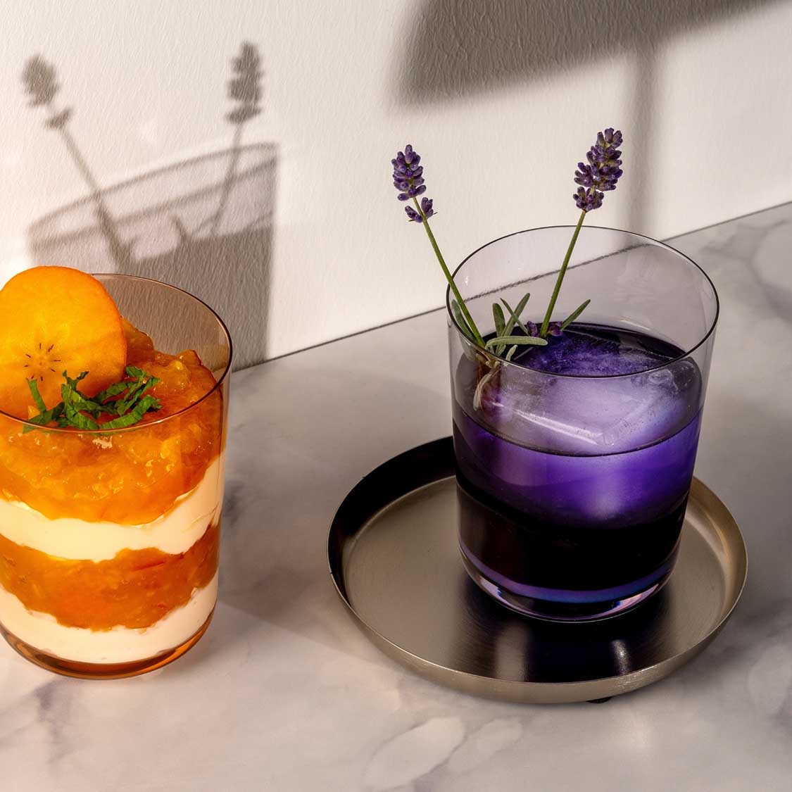 Trinkglas LEGGERO mit lila Getränk, Lavendelblüten, neben Cocktail mit Orangenscheibe