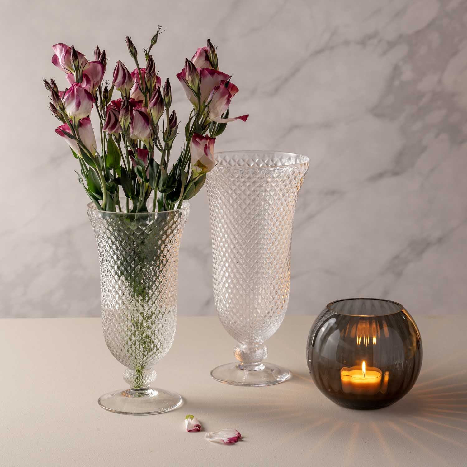 Zwei Glasvasen mit strukturierter Oberflaeche neben brennender Kerze und rosa Nelken in Dekoszene