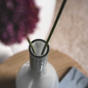 Vase BEAUTY grau mit Blumenstiel und unscharfem Blütenhintergrund auf Holztisch