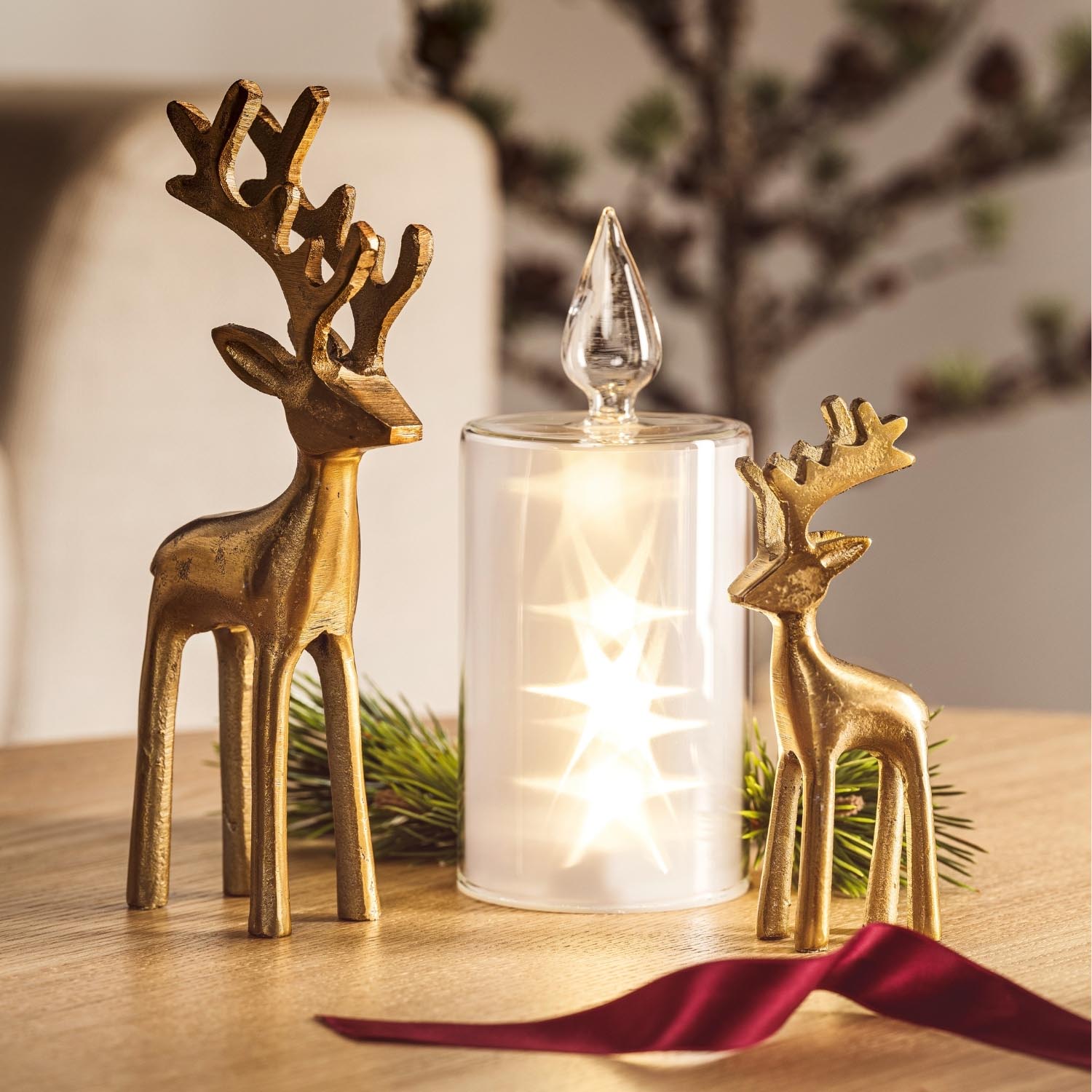 LED Kerze RIPOSO mit goldfarbenen Hirschfiguren und rotem Band auf Holztisch