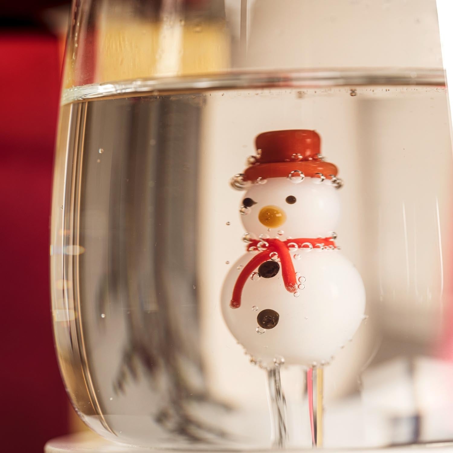 Trinkglas RIPOSO mit Schneemann-Motiv gefüllt mit Wasser, rote Dekoration im Hintergrund