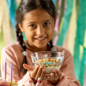Mädchen mit Zöpfen hält Schale BAMBINI AVVENTURA mit Müsli, bunte Wand im Hintergrund