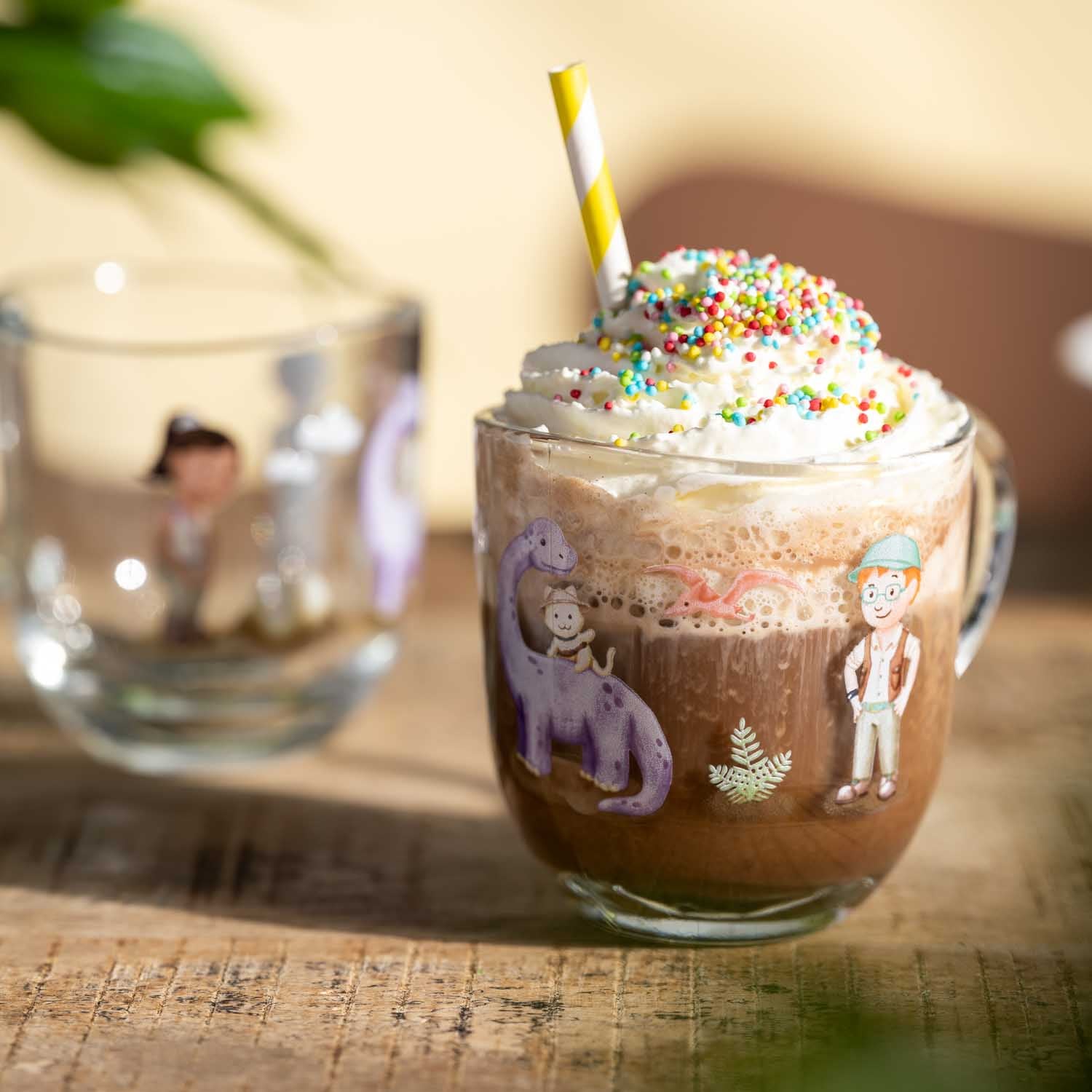 Glastasse BAMBINI AVVENTURA mit bunten Motiven gefüllt mit Kakao, Sahne und Streuseln auf Holztisch