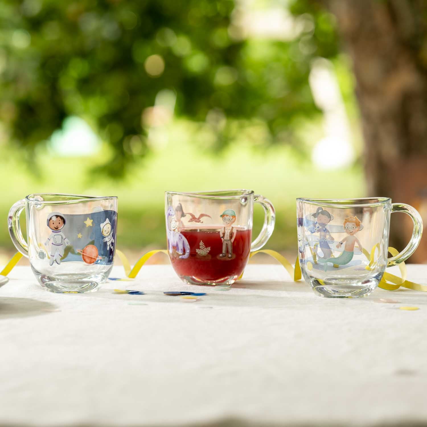 Drei Glastassen mit bunten Kindermotiven auf Tisch im Freien, mittlere Tasse mit rotem Getränk gefüllt