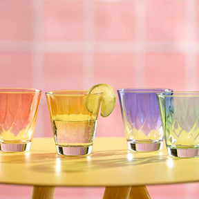 Vier Trinkgläser TWIST mit farbigen Getränken auf gelbem Tisch, rosa Hintergrund, Zitronenscheibe als Garnitur