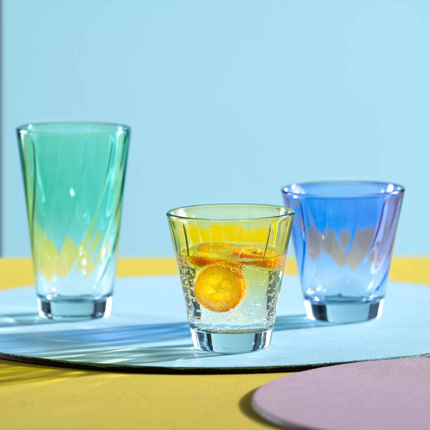 Drei Trinkgläser TWIST in Grün, Gelb und Blau mit Getränk und Zitronenscheibe auf farbigem Untergrund
