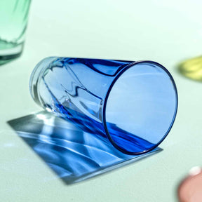 Blaues Trinkglas TWIST 300 ml liegt auf weißer Oberfläche, daneben grüne und rosa Gläser